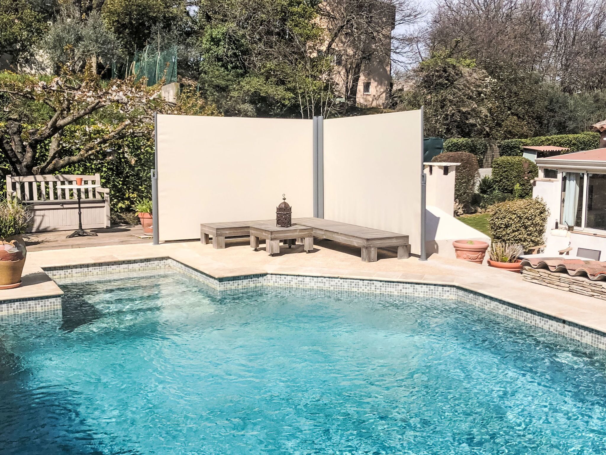 Une piscine dotée d’un banc intégré et d’une petite table, entourée de plantes en pot et d’un écran de protection blanc.