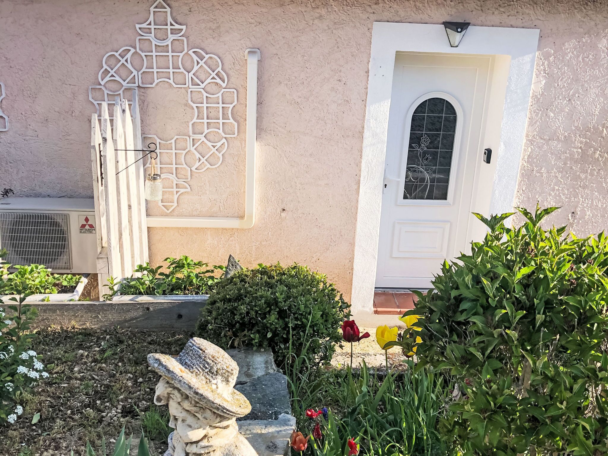 Un edificio con una puerta blanca, un adorno en la pared y un jardín con diversas plantas y flores.