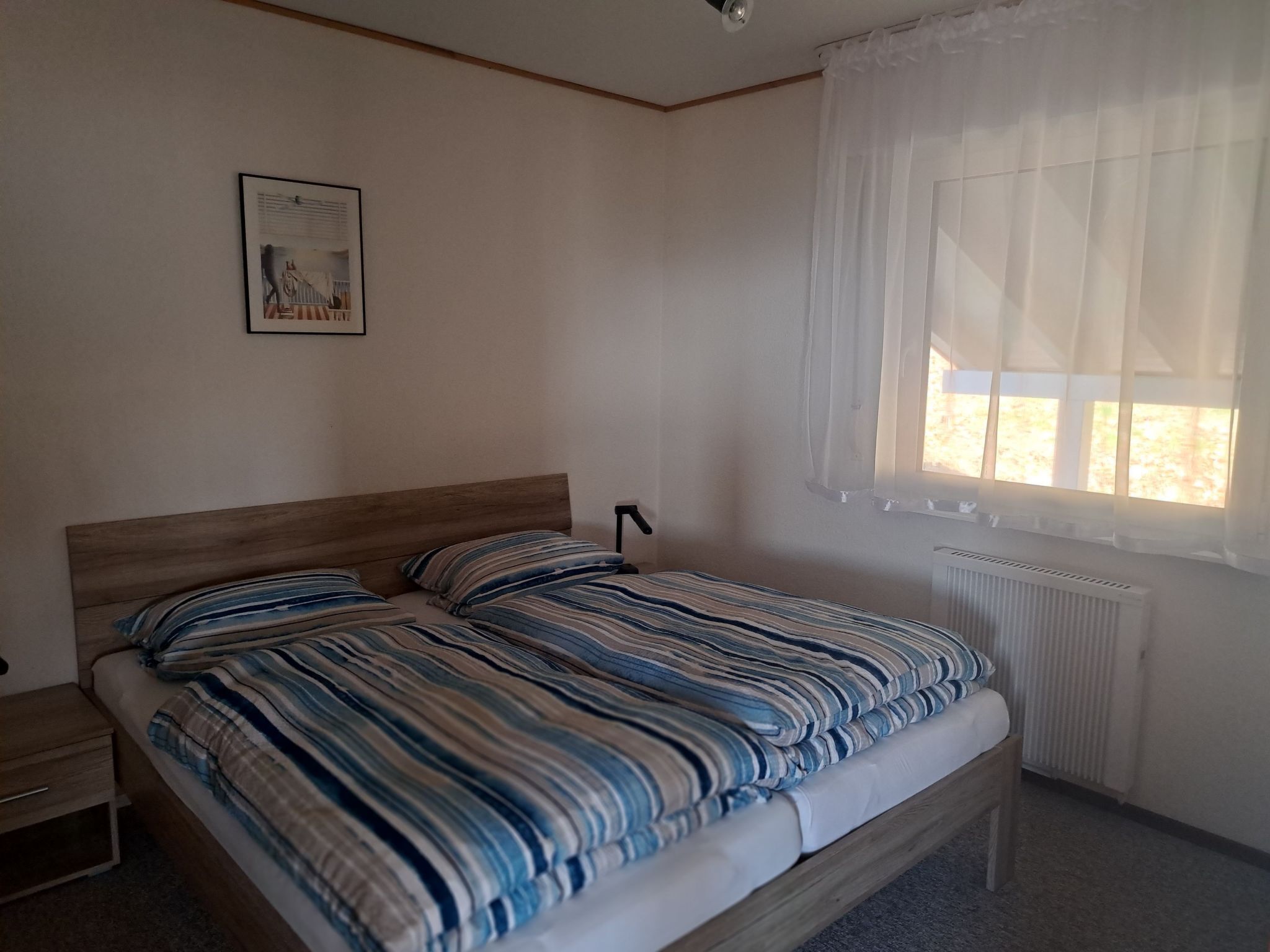 Une chambre à coucher avec un lit en bois, une literie à rayures, une table de chevet, un radiateur et une fenêtre ornée de rideaux légers.