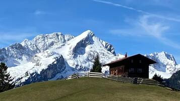 Eine Berglodge mit Holzwänden, umgeben von einem Holzzaun, vor einer Kulisse schneebedeckter Gipfel unter blauem Himmel.