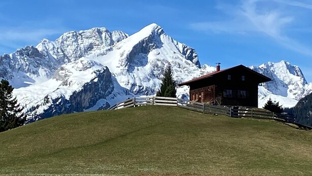 Un chalet de montagne aux murs en bois, entouré d’une clôture en bois, se dressant sur un fond de pics enneigés sous un ciel bleu dégagé.
