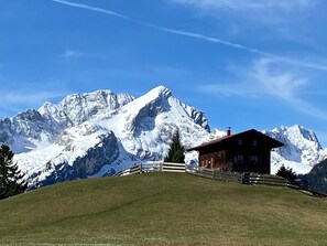 Un chalet de montagne aux murs en bois, entouré d’une clôture en bois, se dressant sur un fond de pics enneigés sous un ciel bleu dégagé.