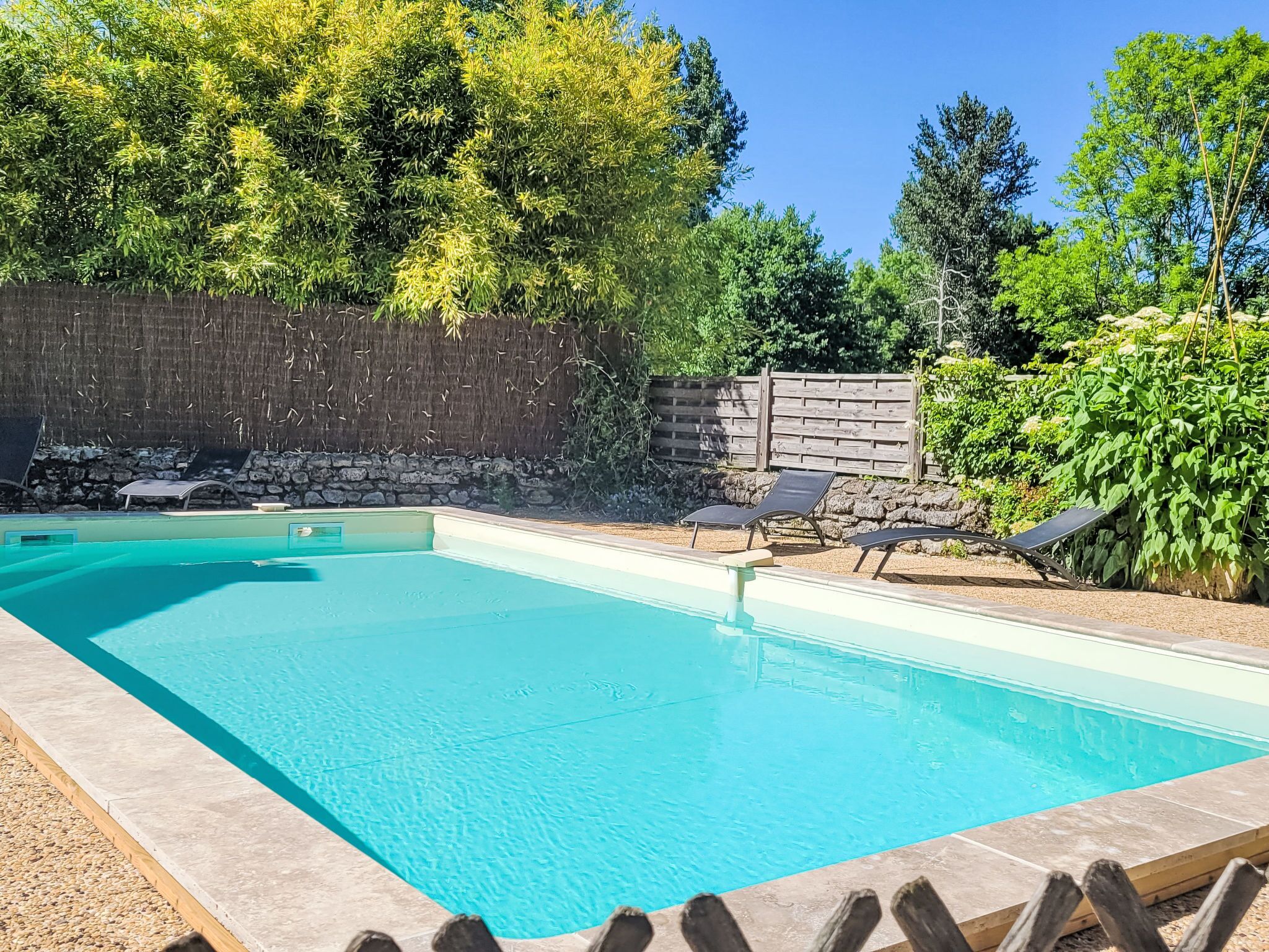 A rectangular swimming pool with a stone border, surrounded by a wooden fence and lush greenery.