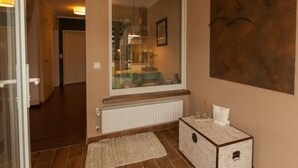 Un pasillo con puerta de vidrio que da a una habitación, un radiador, una alfombra y una pequeña mesa con una caja y una vela.