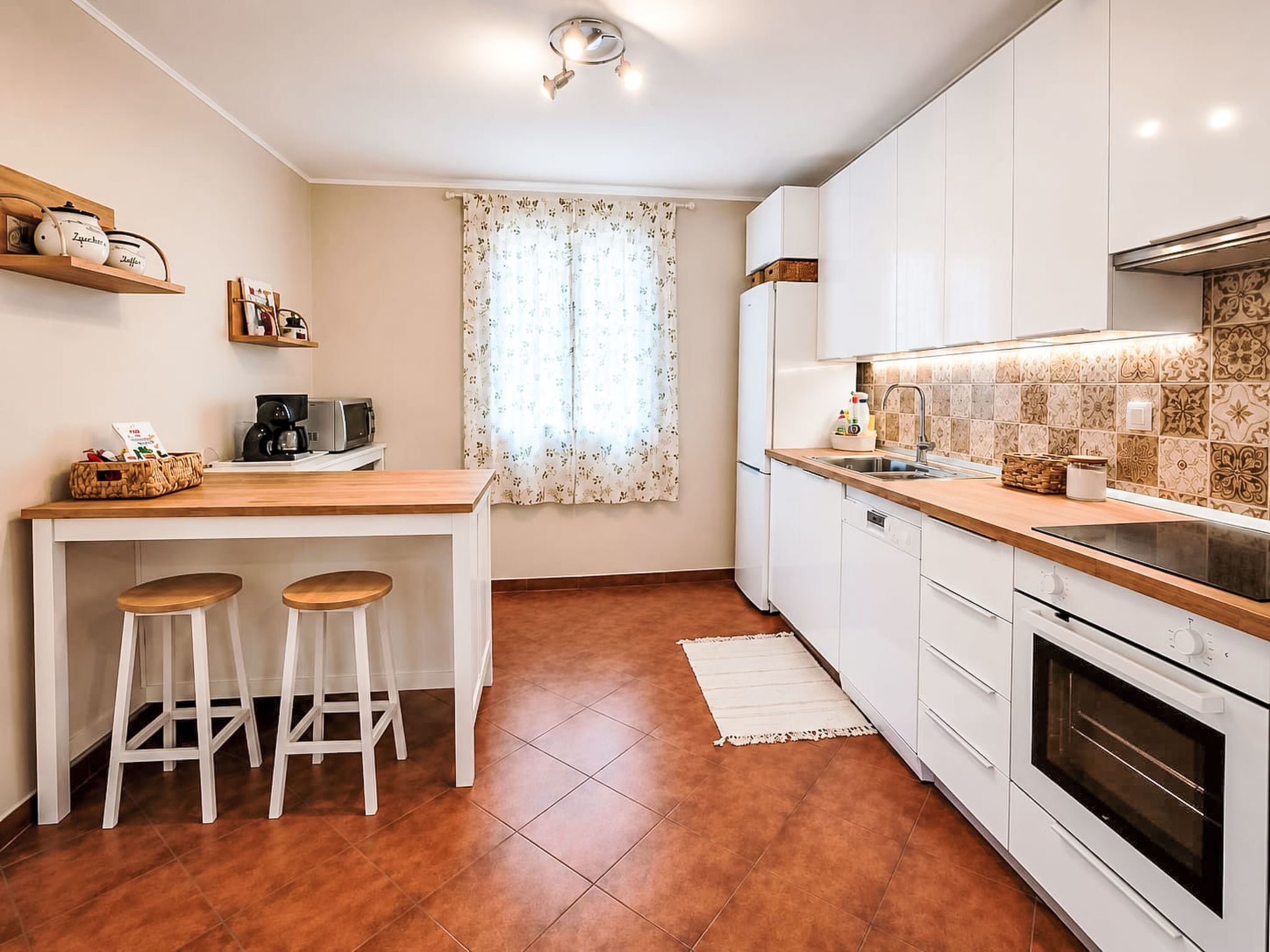 Una cucina moderna con mobili bianchi, un piano di lavoro in legno, due sgabelli, una finestra con tende a fiori e una parete di sopra cucina rivestita di piastrelle.