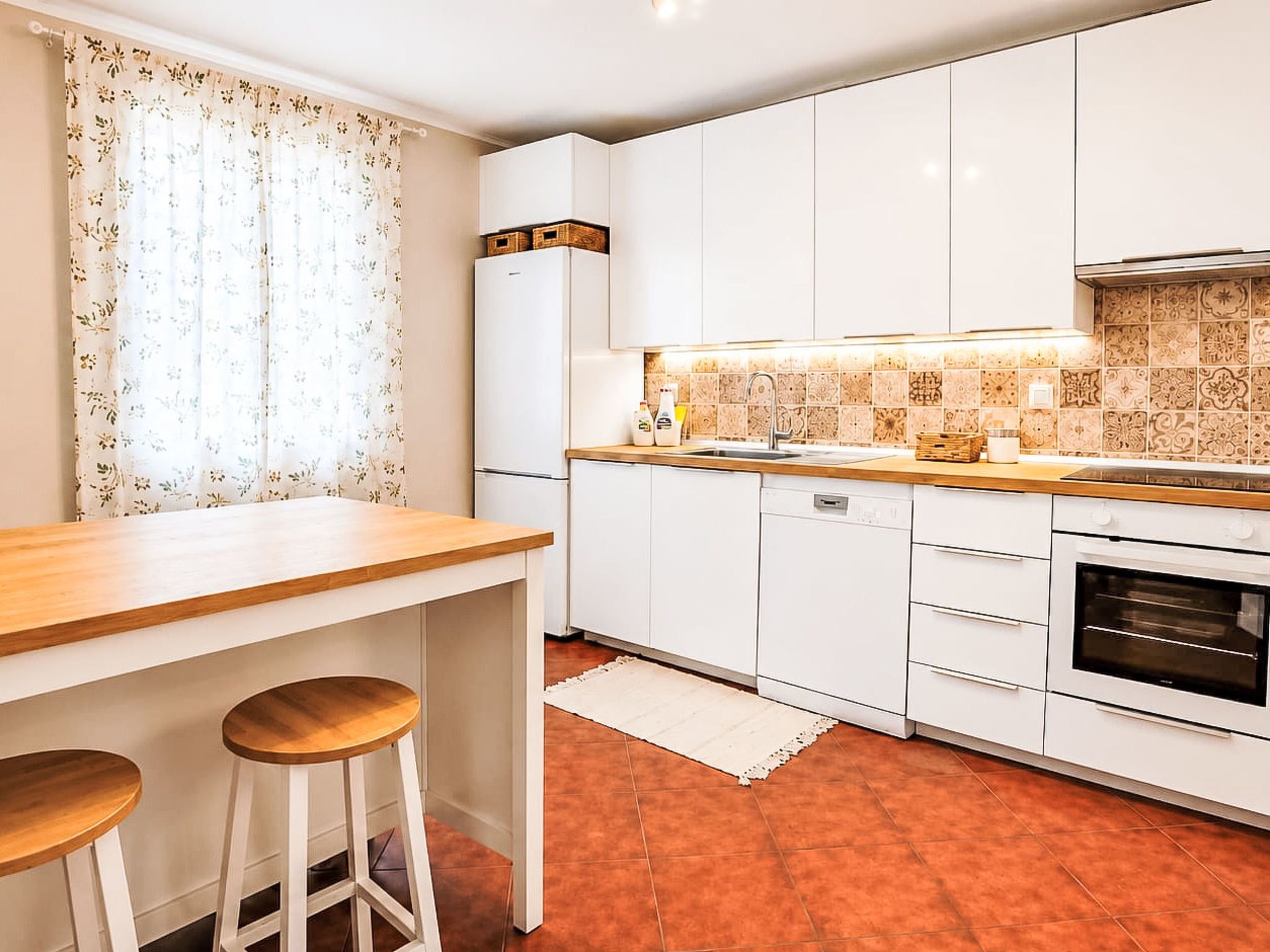 Una cocina con armarios blancos, encimera de madera, horno empotrado y ventana con cortinas florales.