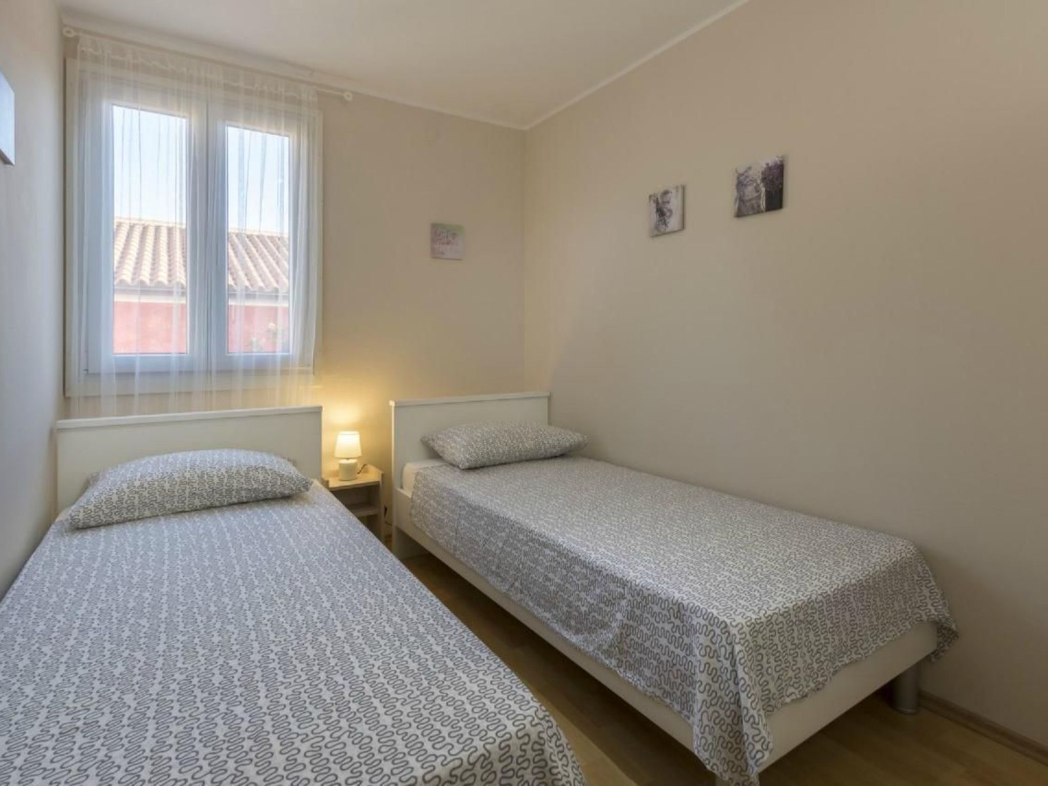 Et lille, rent værelse med to senge, et vindue med let gardin og et lille bord med lampe.