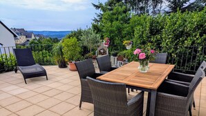 A patio with a wooden table and wicker chairs, surrounded by potted plants and a view of houses in the distance. - Haus Starkenburg by Interhome (Irmenach)