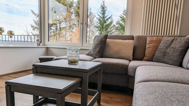 A modern living room with a grey sofa, two dark wooden tables, and large windows offering a view of trees and a clear sky. - Haus Starkenburg by Interhome (Irmenach)