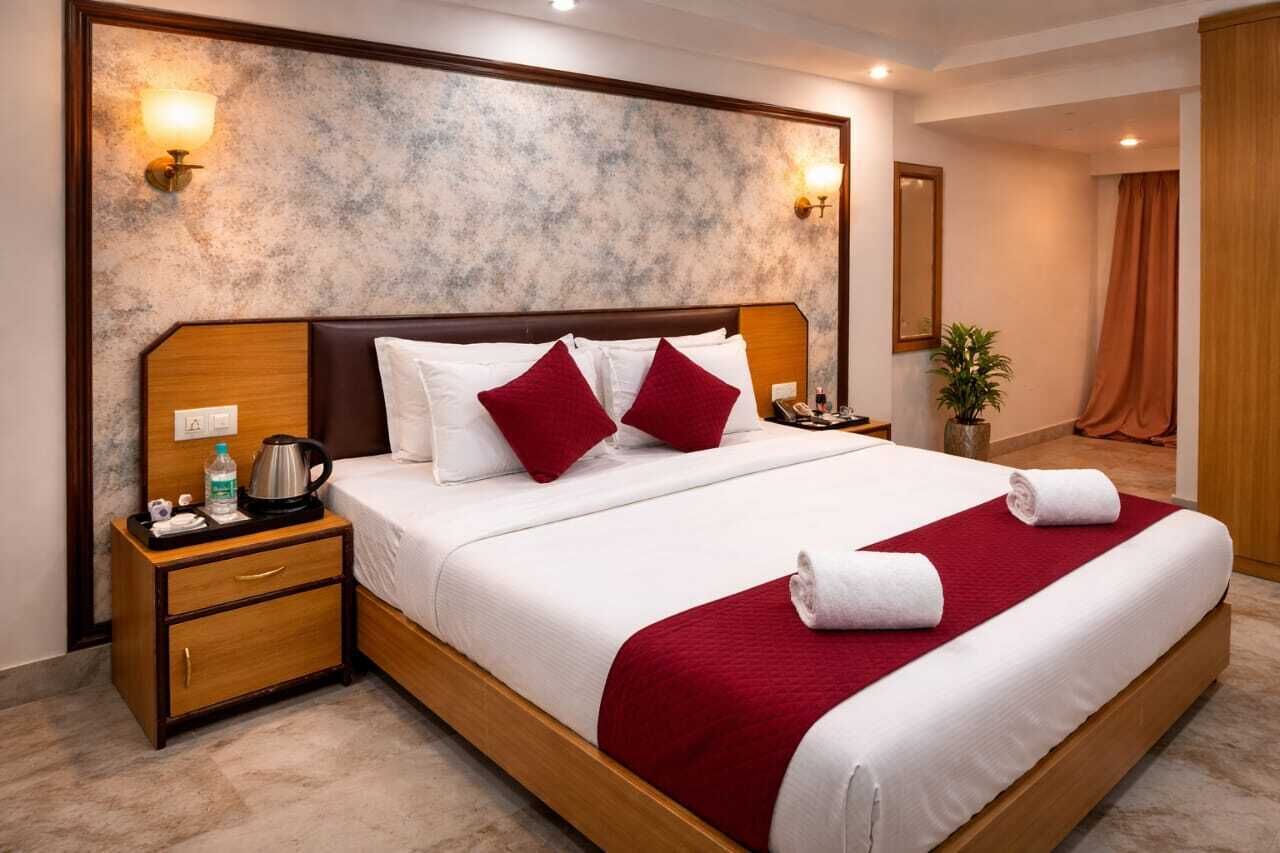 A hotel room with a large bed, wooden headboard, two bedside tables, a nightstand, a red throw blanket, a white pillow, and a wall-mounted light fixture.