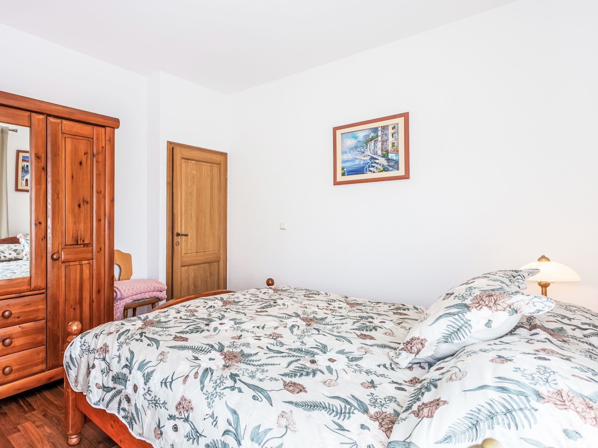 Un dormitorio con un armario de madera, una cama con ropa de cama floral, una puerta de madera y un cuadro enmarcado en la pared.