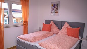 En dobbeltseng med gråt sengegavl og orange sengetøj, et natbord med lampe, og et vindue med gardiner.