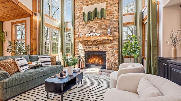 A cozy living room with a stone fireplace, a green sofa, a white armchair, and a patterned rug.