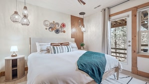 A bedroom with a bed, bedside table, lamp, wall decor, and a view through a door.