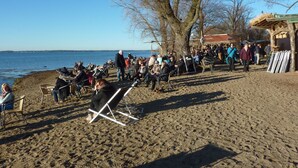 Een strand met mensen die op stoelen zitten en sommigen staan, een grote boom en een gebouw met rieten dak.