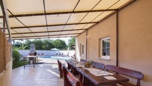 Un patio al aire libre con un área de estar cubierta, una parrilla para barbacoa y una piscina al fondo.