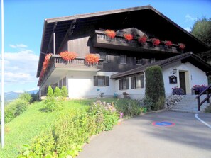 Un chalet de dos pisos con techo inclinado, balcones con jardineras y una entrada principal con escalones.