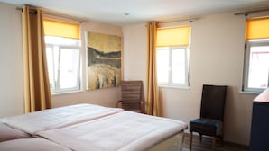 Una cama doble con cabecera, dos ventanas con cortinas, una silla y un cuadro en la pared.
