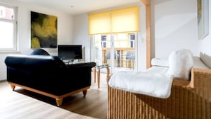 Una sala de estar con un sofá negro, televisión, un cuadro grande y una silla de mimbre.