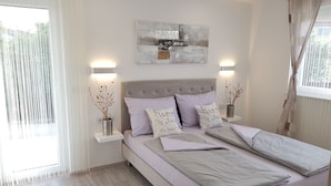 Un dormitorio moderno con una cama grande, dos mesitas de noche con lámparas, un cuadro abstracto en la pared y una ventana con cortinas translúcidas.