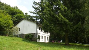 Casa de dos pisos con base de piedra, rodeada de abundante vegetación y una hamaca.