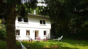 Casa de dos pisos con fachada blanca, una chimenea visible y una puerta principal. Hay dos ventanas en la planta baja y una puerta en el piso superior. Una hamaca está sujeta entre dos árboles en primer plano, y hay dos sillas y una mesa en el patio.