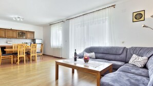 Ampla sala de estar com sofá cinzento, mesa de jantar de madeira e área de cozinha com armários de madeira e painel de azulejos.