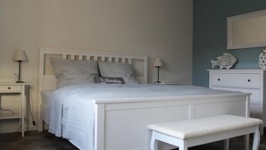 A bedroom with a white bed, two bedside tables with lamps, a bench, and a dresser with decorative fish. - Lüntjenüst by Interhome (Marienhafe)