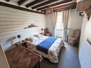 Une chambre à coucher avec un lit en bois, une table de chevet, une chaise, une fenêtre avec des rideaux et un luminaire suspendu.