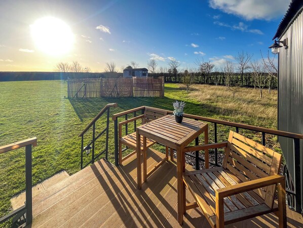 A wooden deck with a table and bench, overlooking a grassy field and a wooden structure in the distance.