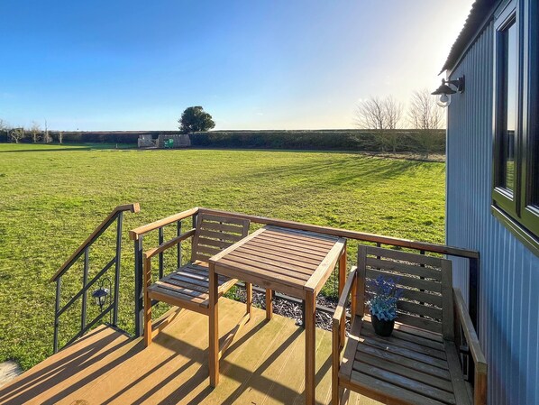 A wooden deck with a table and chairs, overlooking a grassy field and a building with a blue exterior.