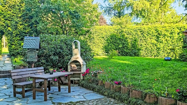 Une terrasse avec une table en pierre et des bancs, entourée d’un jardin agrémenté d’un four à pizza et d’une lanterne.