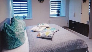 Une chambre à coucher avec un lit, deux fenêtres équipées de stores, une armoire et un miroir.