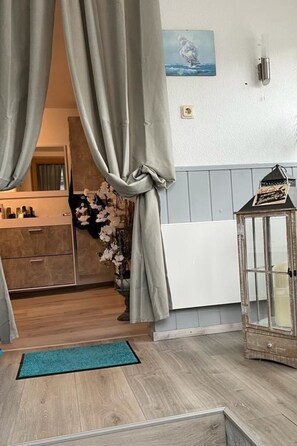 Una habitación con piso de madera, un tapete azul, un radiador blanco, una lámpara de pared, un cuadro enmarcado y un pequeño armario de madera.