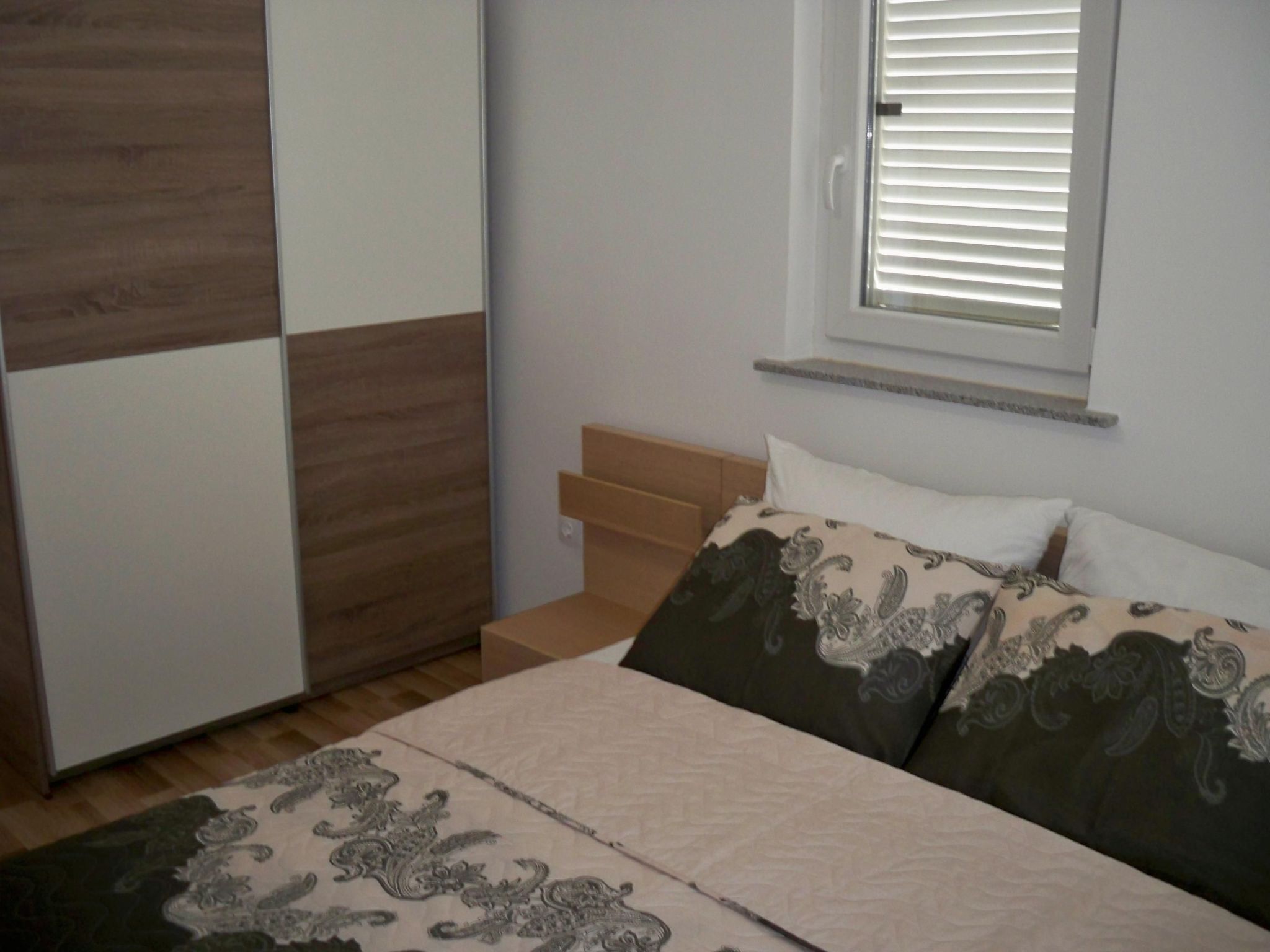 A bedroom with a bed, a window with blinds, a wooden headboard, and a patterned rug.