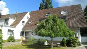 Ein zweigeschossiges Haus mit einem braunen Ziegeldach, weißen Wänden und einem Vorgarten mit einem großen Baum.