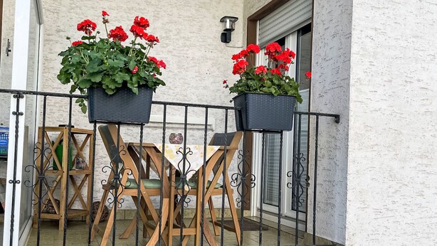 Ein Balkon mit zwei Kübeln roter Blumen, einem Holzstuhl und einem Tisch.
