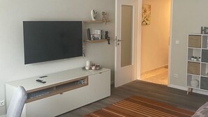 Ein modernes Schlafzimmer mit einem Flachbildfernseher, einer weißen Ablage, einem Streifen Teppich und einem Bett mit grau-weißer Musterdecke.
