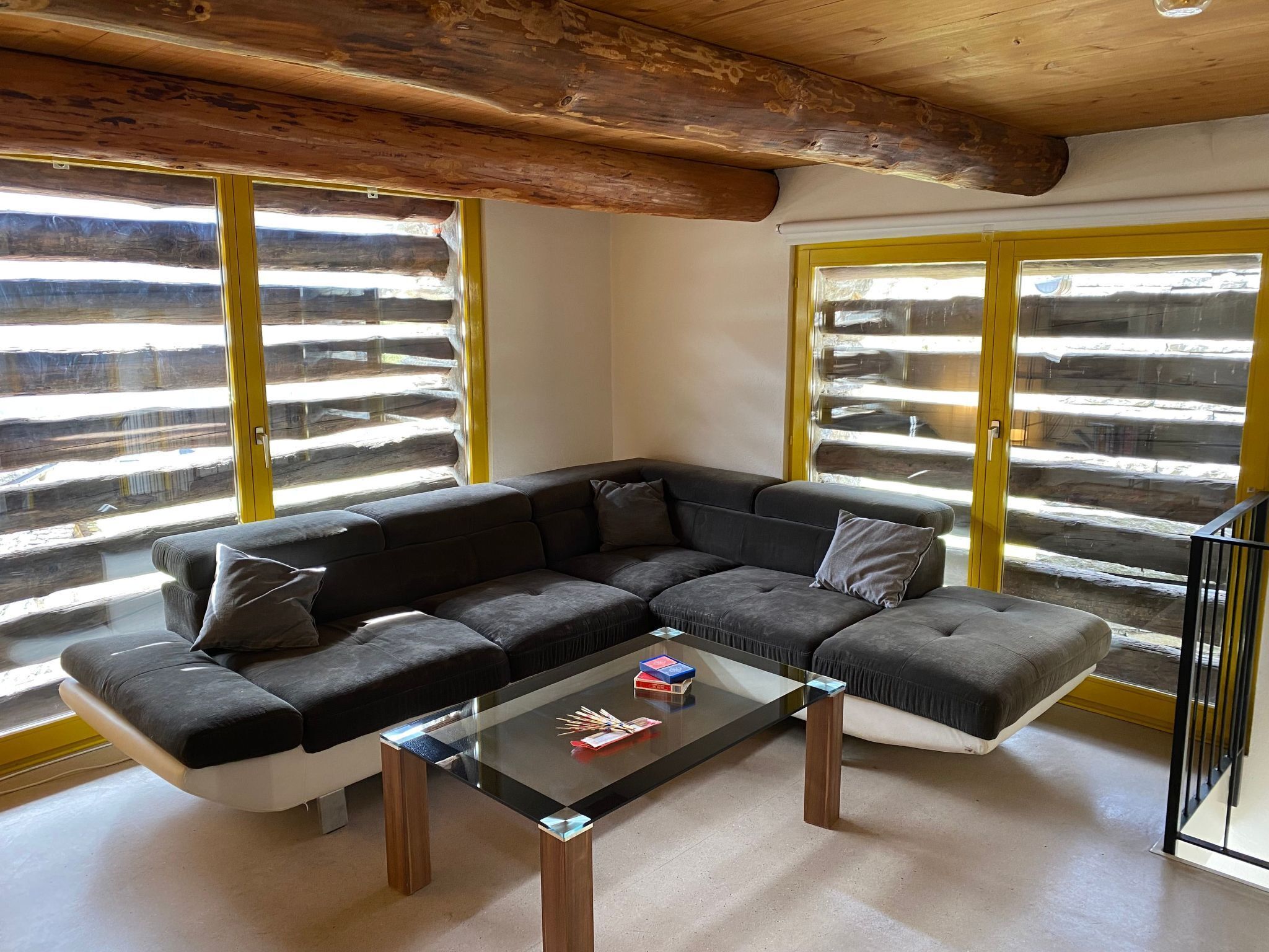 Ein modernes Wohnzimmer mit einem Ecksofa, einem Glastisch und großen Fenstern mit Holzrahmen.