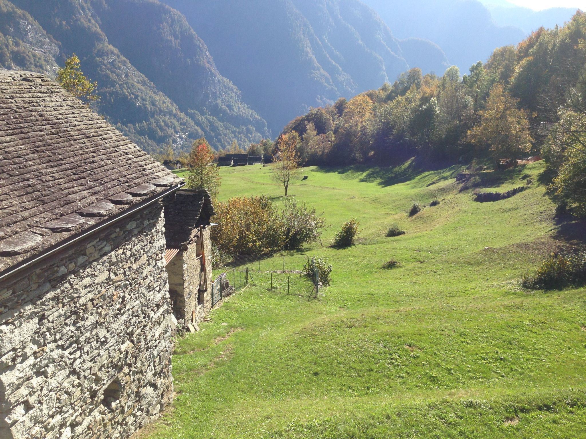 Ein Steinhaus mit geneigtem Dach, das auf ein grünes Tal mit Bäumen und Bergen im Hintergrund blickt.