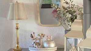 Eine dunkle Holzkommode mit einer Goldlampe, einem Spiegel und Dekorationselementen, darunter eine Vase mit Blumen und eine kleine Figur.