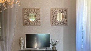 Ein Wohnzimmer mit Flachbildfernseher auf einer Holzkommode, zwei Zierteilen, einer Kristallleuchtreiter und weißen Möbeln.