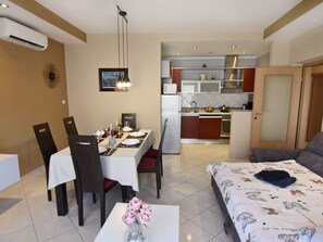 Uma sala de jantar com uma mesa posta para quatro pessoas, uma cozinha ao fundo e uma cama com colcha estampada.