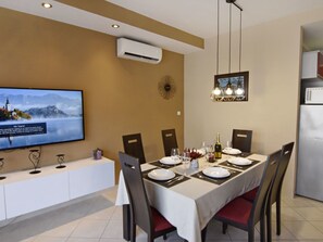 Uma área de refeições com uma mesa posta para quatro pessoas, uma televisão fixada na parede e um aparelho de ar condicionado sobre a mesa.