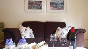 Een woonkamer met een donkerbruine bank, bloemenkussens, een witte tafel met een boek, twee wijnglazen, een fles wijn en twee vazen met bloemen.