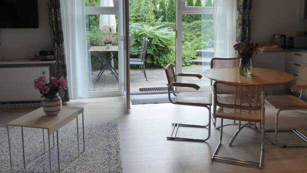 Een moderne woonkamer met een grote schuifpui naar een tuin. Er is een eethoek met een ronde tafel en stoelen, een klein keukentje en een flatscreen tv aan de muur.
