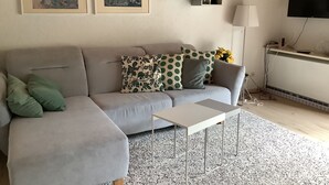 Een woonkamer met een grijze bank, een witte bijzettafel, een decoratief kussen en twee ingelijste kunstwerken aan de muur.