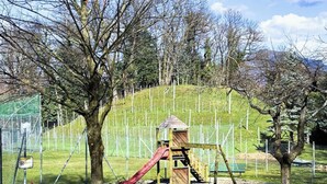 Un parque infantil con tobogán y estructuras para escalar, rodeado de árboles y una zona vallada.
