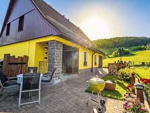 Una casa de fachada amarilla, chimenea de piedra y patio con una parrilla y sillas.