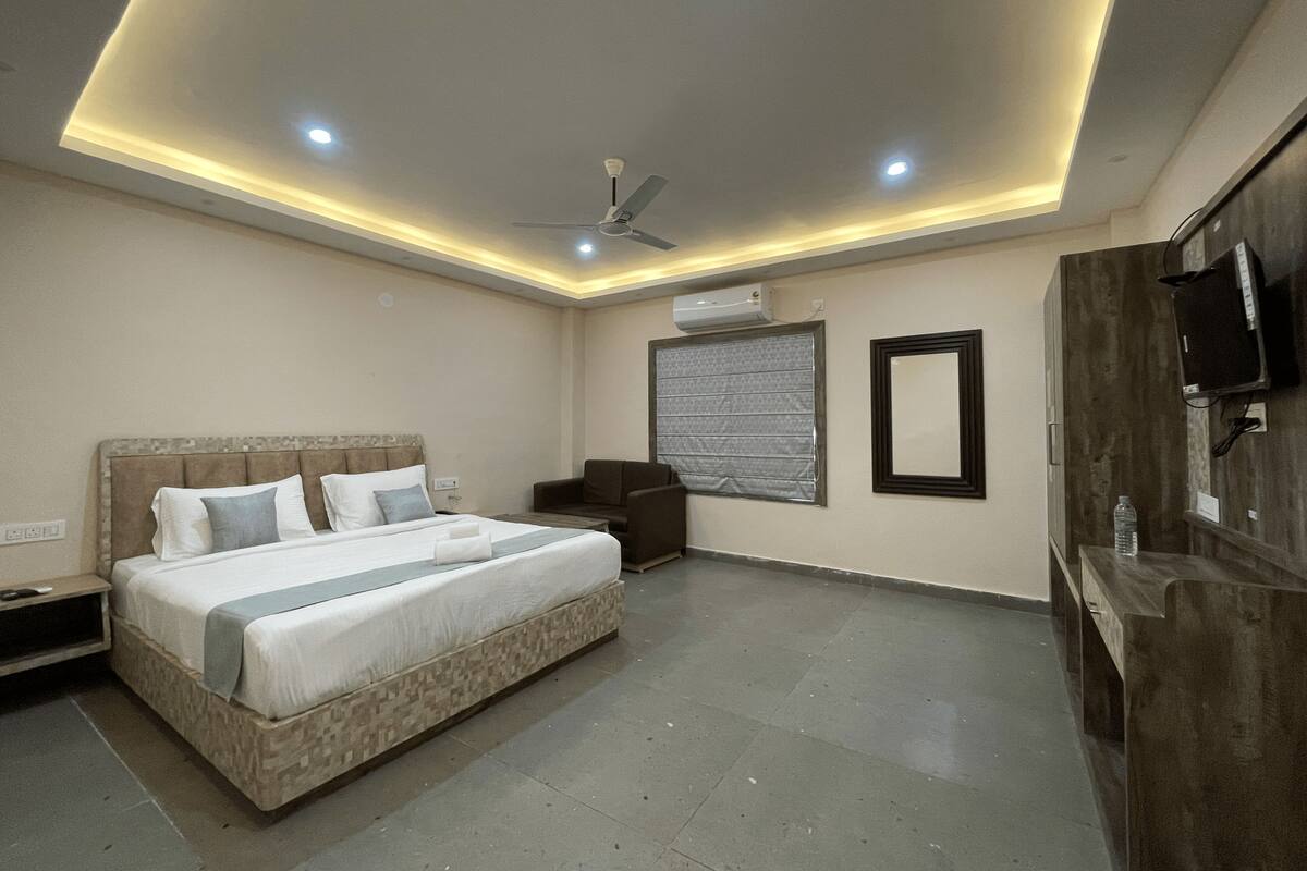 A modern hotel room with a large bed, a ceiling fan, recessed lighting, a window with blinds, and a wooden wardrobe.